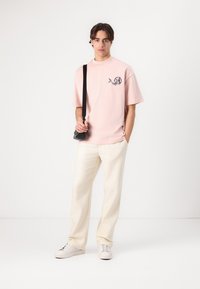 Pink cotton t-shirt with a graphic design, paired with off-white trousers and white sneakers. Model holds a black crossbody bag.