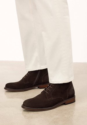 Botas de ante marrón hasta el tobillo con puntera redondeada, cordones en la parte delantera y un tacón bajo, combinadas con pantalones de color claro. Los laterales tienen detalles de cuero liso.