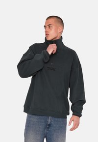 Sweatshirt en polaire zippé de couleur vert foncé avec col haut, coupe décontractée et détail de logo sur le devant. Tissu texturé avec coutures visibles.