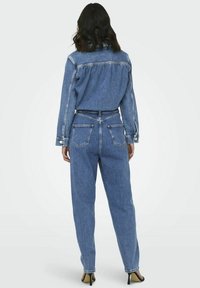 Femme aux cheveux bruns mi-longs portant une veste en jean bleue et un jean taille haute, debout, dos face au spectateur.
