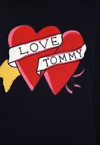 Rotes besticktes Herzdesign mit einem weißen Band, auf dem "LIEBE TOMMY" steht, vor einem dunklen marineblauen Hintergrund. Mit gelben Akzenten.