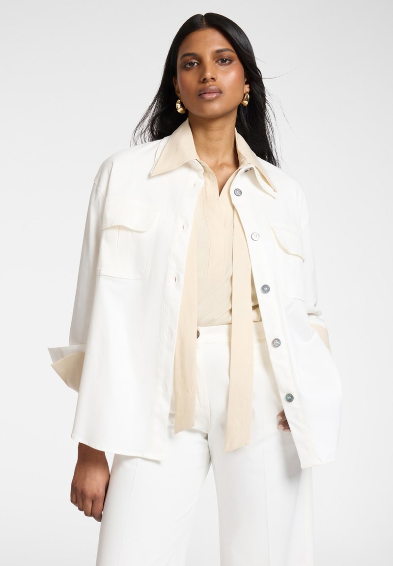 Donna con lunghi capelli neri che indossa una giacca bianca oversize abbottonata, una camicetta beige con lunghe coulisse, pantaloni bianchi e orecchini a cerchio dorati, in piedi con sicurezza.