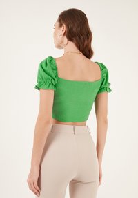 Top corto verde con maniche a sbuffo, tessuto testurizzato, scollatura quadrata; abbinato a pantaloni beige a vita alta con doppie asole per la cintura.