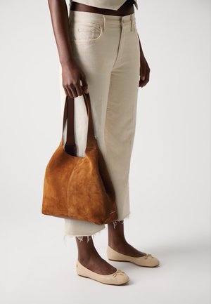 Sac en cuir suédé marron avec une texture douce et une forme décontractée, porté par une personne en pantalon clair et des ballerines.