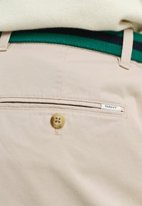 Beige broek met een achterzak, een crème knoop en een groen gestreepte geweven riem met donkerblauwe accenten. GANT-logo zichtbaar op de zak.