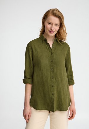 Frau mit heller Haut und roten Haaren, die ein olivgrünes Hemd mit hochgekrempelten Ärmeln und beige Hosen trägt, steht vor weißem Hintergrund.