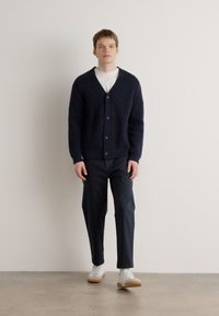 Ναυτικό πλεκτό cardigan με λαιμό V και κουμπιά, συνδυασμένο με σκούρους παντελόνες και λευκά αθλητικά παπούτσια. Λείο υλικό με ραβδωτό μοτίβο.