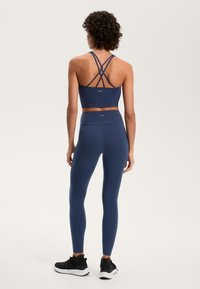 Kobieta w granatowym stanik sportowy z krzyżującymi się ramiączkami i pasujących wysokich legginsach, stojąca tyłem na jednolitym tle.