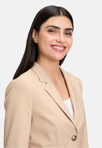 Beżowa marynarka z wyraźnymi klapami i zapięciem na jeden guzik. Gładka faktura materiału, dopasowany krój i proste detale projektowe.