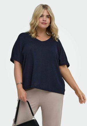 Lyshåret kvinde iført en navy kortærmet top og beige bukser, der holder en sort håndtaske med én hånd mod en ensfarvet baggrund.