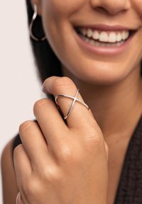 Silberner Crossover-Ring, der am Finger getragen wird, mit einer glatten, glänzenden Oberfläche und einem minimalistischen Design. Hand und Gesicht teilweise sichtbar.