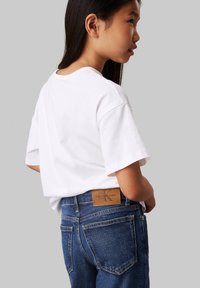 White cotton t-shirt with short sleeves, paired with blue denim jeans. Notable details include a brown leather waistband label.