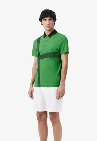 Grön pikétröja med svarta detaljer, kort ärm och tre knappar. Har ett texturerat svart sprutmönster. Bärs med vita shorts.