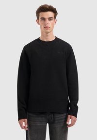 Zwarte gebreide sweater met een V-hals en ribbelzoom. Heeft lange mouwen en een textuurafwerking. Draagt over donkerblauwe denim jeans.