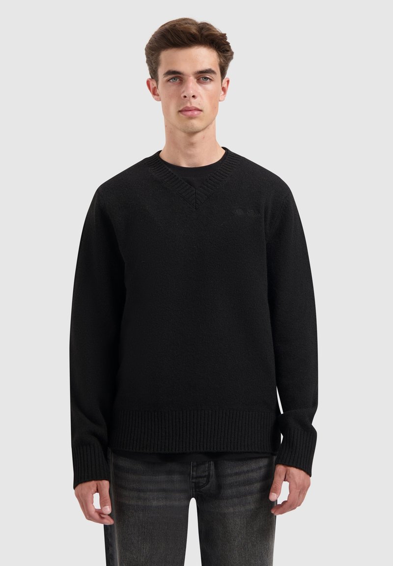 Zwarte gebreide sweater met een V-hals en ribbelzoom. Heeft lange mouwen en een textuurafwerking. Draagt over donkerblauwe denim jeans.