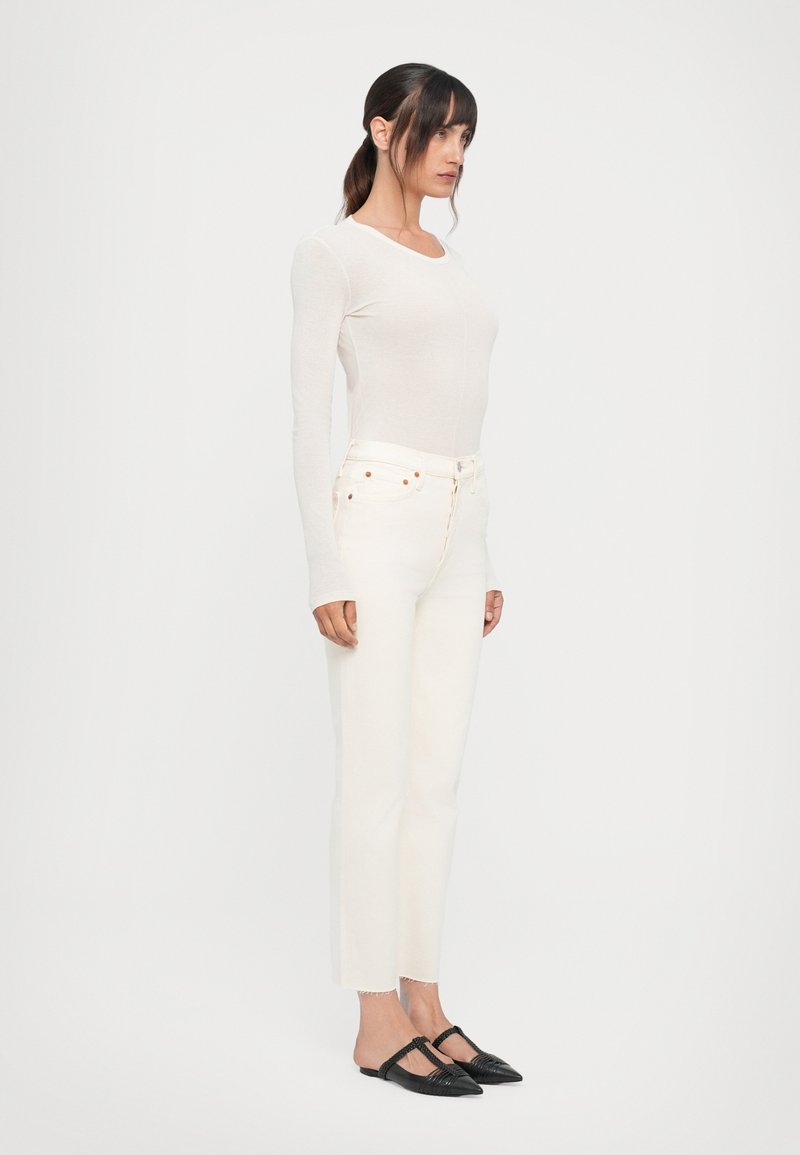 Haut à manches longues clair et jean blanc cropped avec ourlet effiloché ; chaussures plates noires avec détail tressé. Design simple et ajusté.