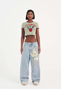 Geknipte olijfgroene t-shirt met rode afwerking en appelafbeelding; lichtblauwe wijde jeans met vlinderborduursel en delicate patronen.