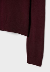 Manche et ourlet d'un pull en maille à manches longues de couleur bordeaux foncé avec poignets et taille côtelés sur un fond clair uni.