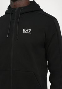 Črna zip-up kapuca iz mehkega materiala. Ima sprednji žep, nastavljive vrvice in belo EA7 logo na prsih.