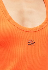 Csíkozott narancssárga ujjatlan felső, amelynek lekerekített kivágása és ezüst színű logói díszítése van a középső részen. Az anyag sima tapintású.