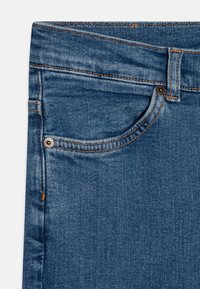 Jeans i middels blå med et lett falmet mønster, som har en frontlomme, metallknapp og oransje søm-detaljer.