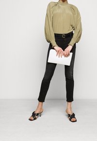 Blusa de manga larga verde oliva con detalles plisados, jeans skinny negros, sandalias de tacón negras con un lazo y un clutch blanco.