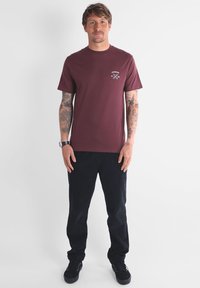 Homme avec des tatouages portant un t-shirt bordeaux et un pantalon noir, se tenant devant un fond blanc uni.