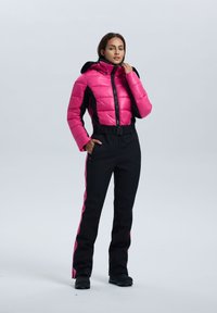 Chaqueta rosa acolchada con detalles negros, cuello alto y cremallera. Pantalones negros con franjas laterales rosas y bolsillos. Calzado negro completa el conjunto.