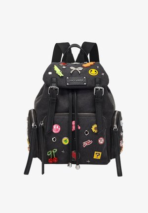 Zaino nero con tasche frontali e laterali con cerniera, decorato con spillette colorate a tema cartone animato, inclusi volti sorridenti, ciliegie, girasole e vari simboli.