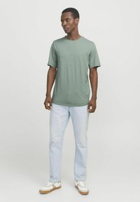 Green short-sleeve t-shirt in a slim fit, paired with light blue straight-leg jeans and white sneakers with tan soles.