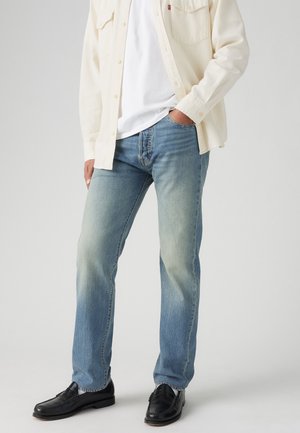 Homme portant un jean bleu clair délavé, des chaussures noires, un t-shirt blanc et une chemise crème boutonnée, main dans la poche, debout devant un fond uni.