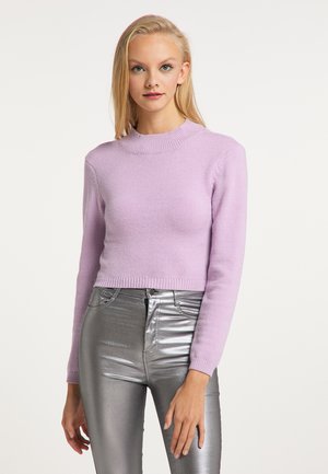 Blondynka ubrana w jasnofioletowy dopasowany sweter i błyszczące srebrne spodnie z wysokim stanem, stojąca na jednolitym tle.