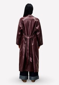 Person mit langen schwarzen Haaren, die einen glänzenden burgunderfarbenen Trenchcoat, weit geschnittene dunkle Hosen und braune Schuhe trägt und mit dem Rücken zum Betrachter steht.