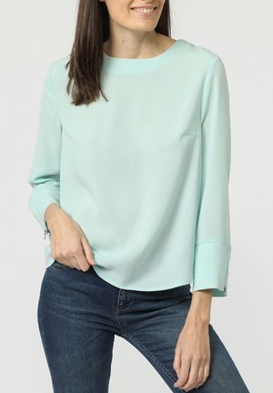 Blusa verde menta hecha de tejido suave, con escote redondo, mangas largas y sutiles detalles de cremallera en los puños.