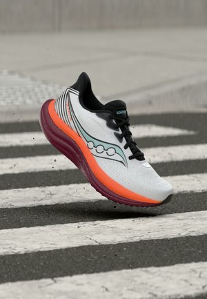 White and black running shoe with orange and red sole mid-air above a striped crosswalk on asphalt surface.