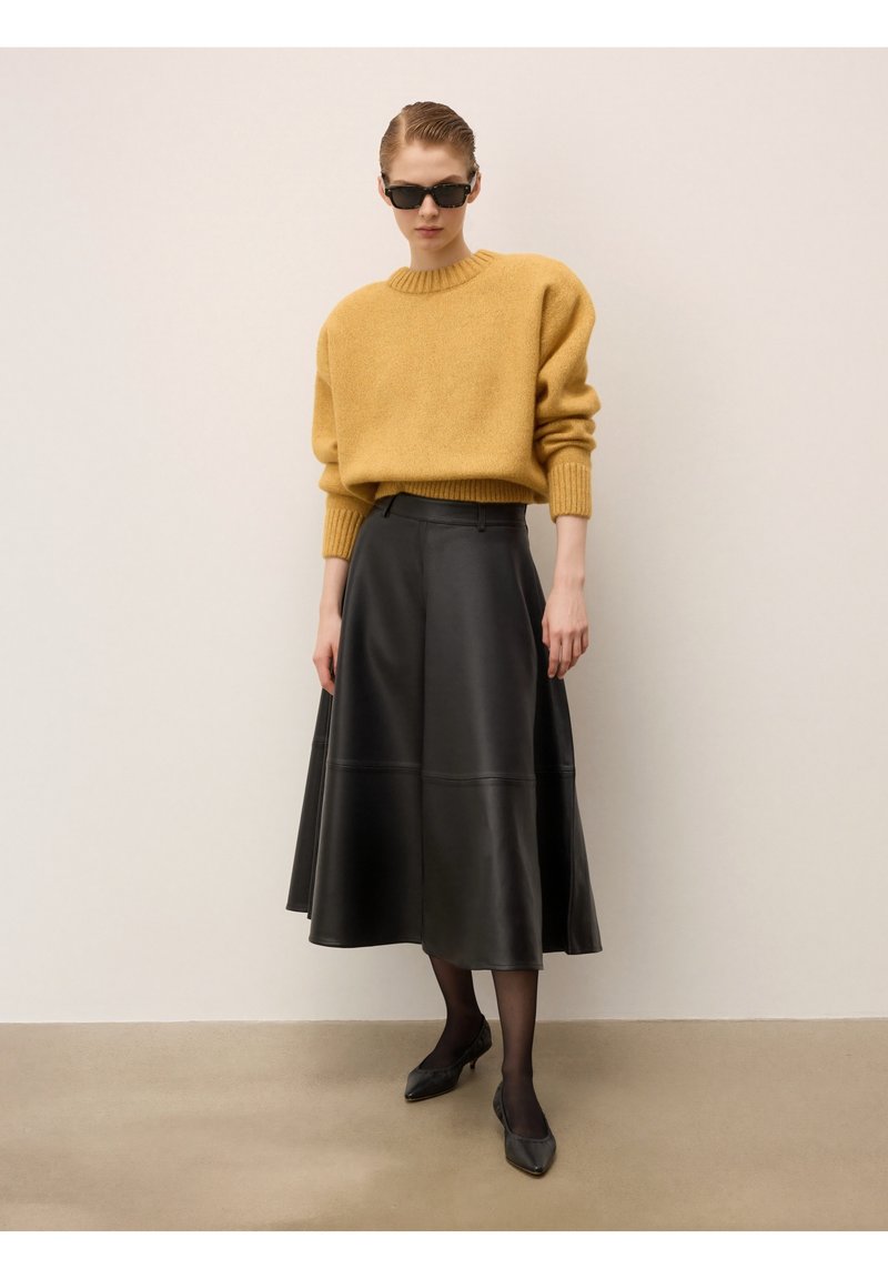 Pull en tricot oversized moutarde, jupe en cuir noir, ballerines noires et lunettes de soleil. Texture lisse, coupe décontractée et silhouette arrivant au genou.