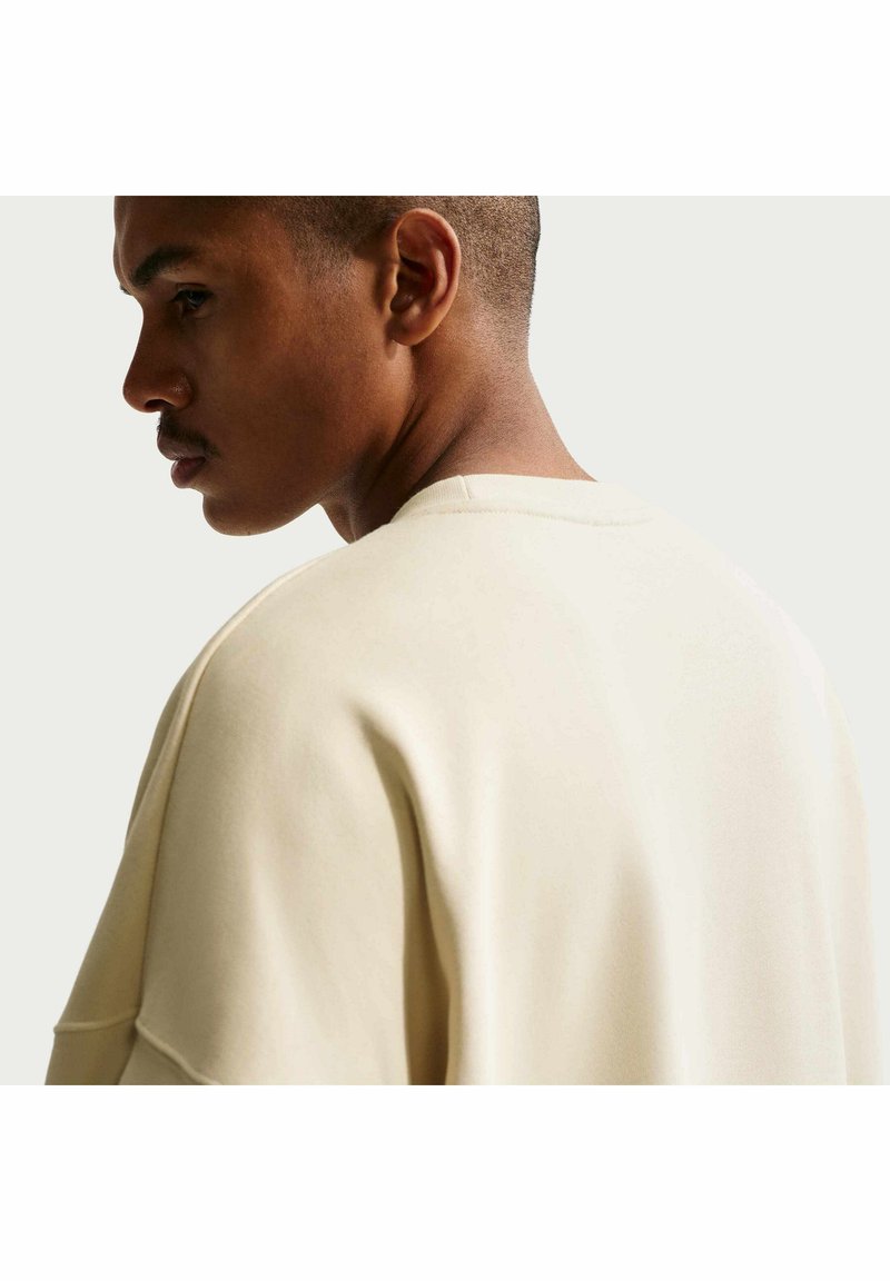 Homme aux cheveux courts portant un pull ample beige clair, vu de côté sur un fond clair et uni.
