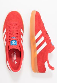 Röda mockasneakers med tre vita ränder, blå logotag och genomskinlig gummisula. Rund tå och texturerad ovandel.