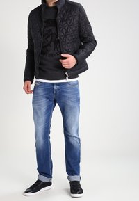 Svart quiltad jacka med hög krage över en mörk tröja, i kombination med ljusblå denimjeans och svarta sneakers med vita sulor.