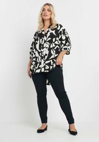 Blusa geometrica bianca e nera con un taglio morbido, maniche a metà, e un orlo alto-basso, abbinata a pantaloni neri slim-fit e scarpe flat.