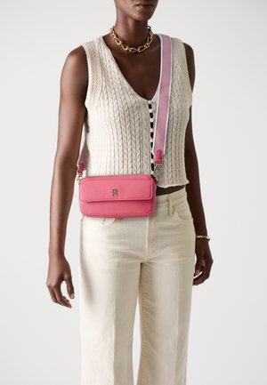 Pink leather crossbody bag with a contrasting strap, featuring a smooth texture and a gold logo accent on the front.