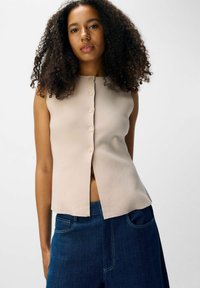 Beige ärmlös topp med rund halsringning, knäppning fram och en slits i nederkanten, kombinerad med mörkblå denimbyxor.