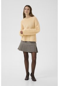 Pfirsichfarbener, flauschiger Pullover mit übergroßen Ärmeln, kombiniert mit einem grauen, karierten, plissierten Rock und dunklen Strumpfhosen, abgerundet mit lila Absätzen.