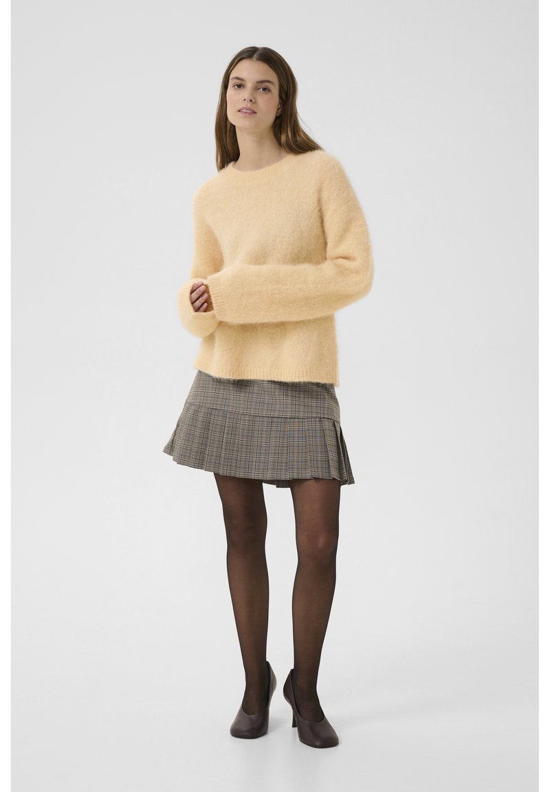 Pull en peluche rose avec des manches surdimensionnées, associé à une jupe plissée à carreaux gris et des collants noirs, le tout complété par des talons violets.