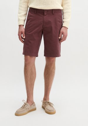 Mann trägt burgunderrote knielange Shorts, cremefarbenen Strickpullover und beige Schnürschuhe, steht vor weißem Hintergrund.