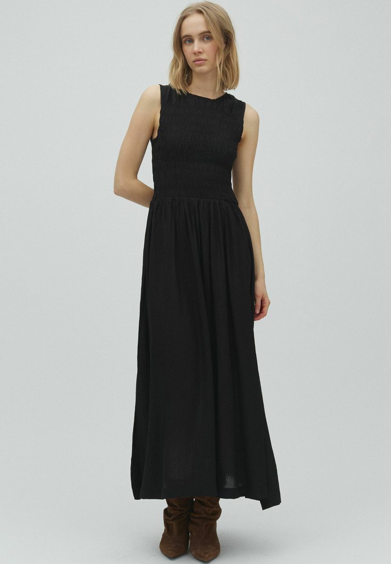 Jeune femme aux cheveux blonds portant une robe maxi noire sans manches et des bottes marron décontractées, debout devant un fond gris uni.