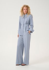 Camicia azzurra chiara a maniche lunghe con colletto e design plissettato. Abbinata a pantaloni a gamba larga, entrambi in tessuto liscio.