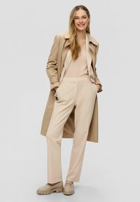 Manteau trench beige sur un haut beige clair, associé à un pantalon beige clair et des mocassins épais assortis. Texture lisse, design minimaliste.