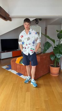 Un jeune homme se tient dans le salon avec une chemise à motifs, un short bleu marine et des baskets bleues à côté d'un canapé marron et une grande plante en pot.
