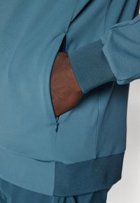 Jachetă sport teal cu buzunar lateral cu fermoar și manșete ribbate. Textură netedă cu un design fără cusături pentru ușurința mișcării.